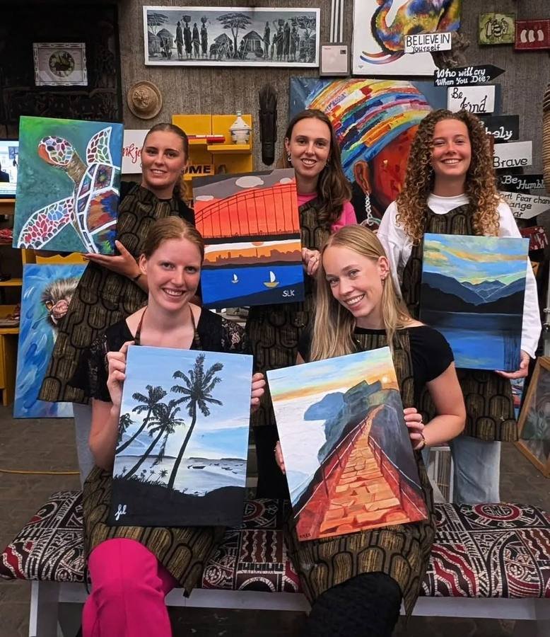 Five visitors holding up the canvases they painted, showing turtles, palms, mountains and a bridge