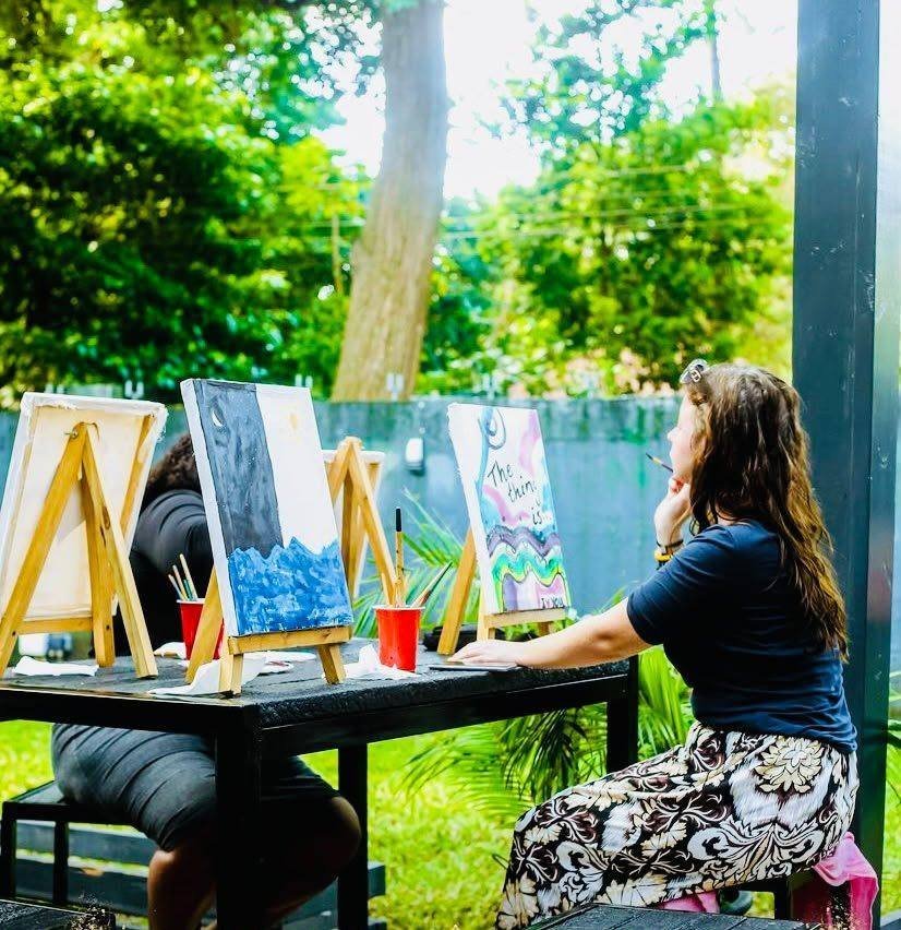 A woman painting at an easel outdoors with green trees behind her