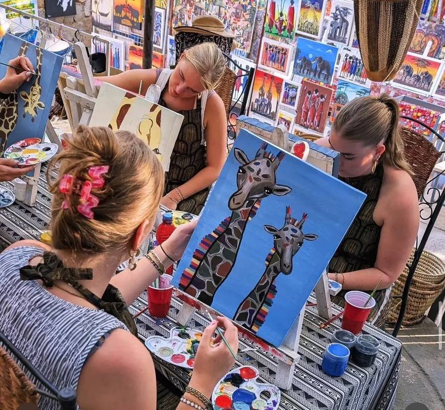 Visitors seated around a table painting giraffes on canvas, with paint palettes and brushes