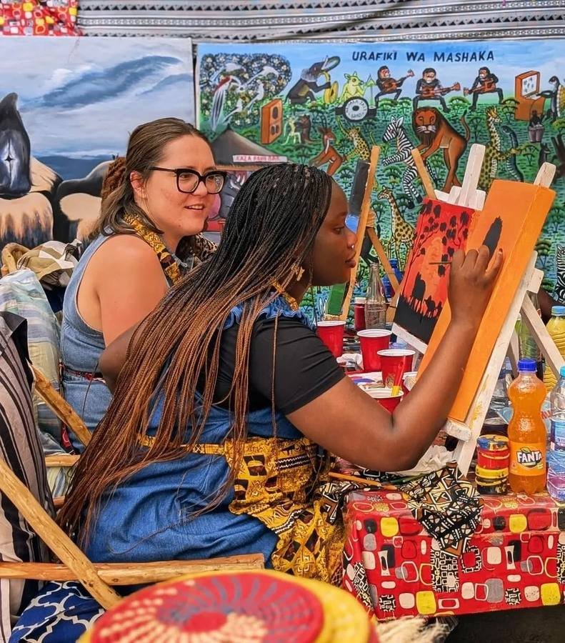 A woman painting an orange savannah sunset with acacia silhouettes while another watches