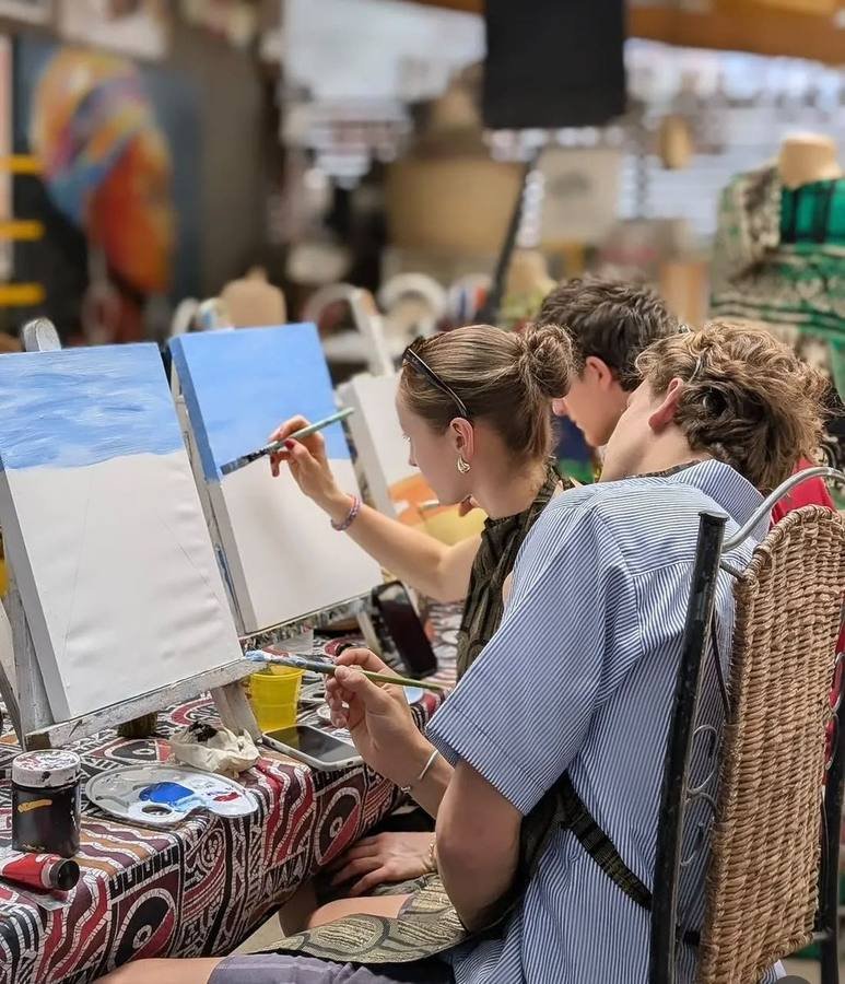 Three visitors painting blue skies on canvases side by side at the workshop