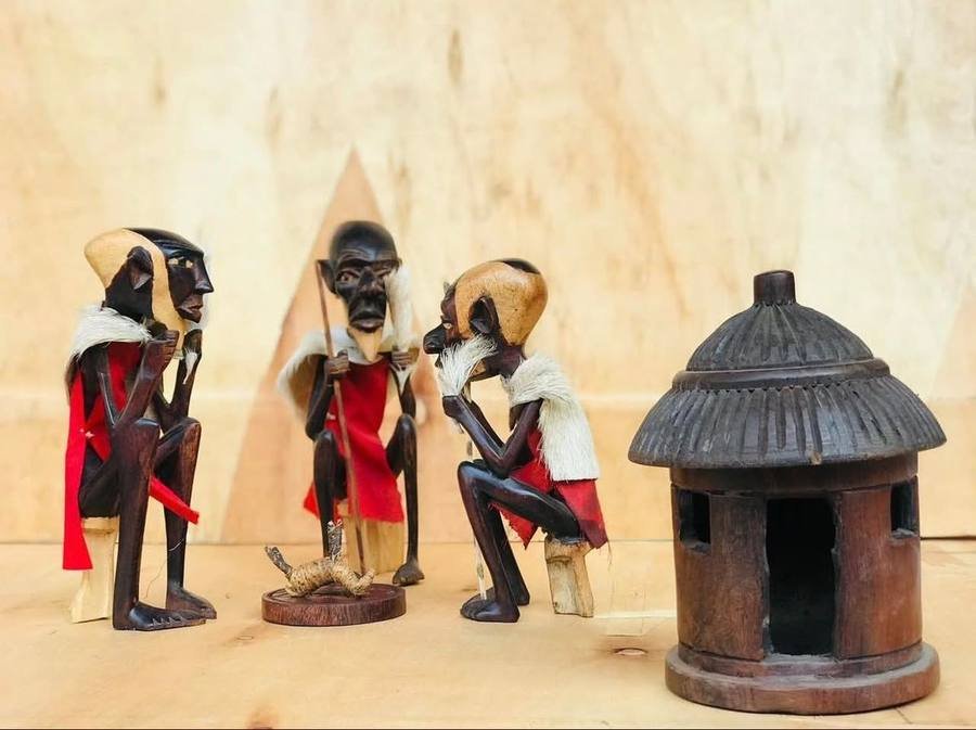 Three carved Maasai elder figurines in red cloth beside a carved wooden hut