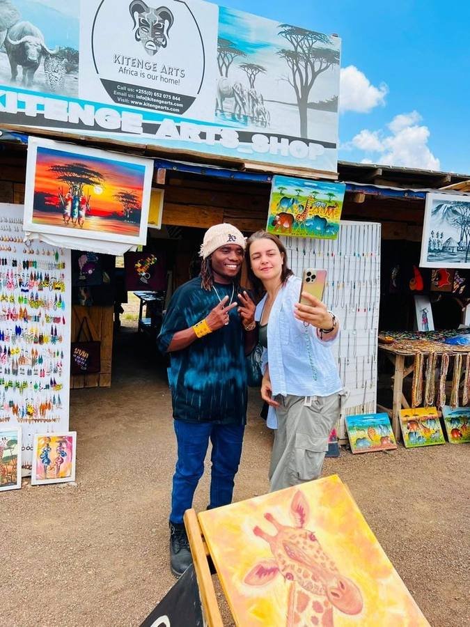 Omary standing with a visitor outside the Kitenge Arts shop, surrounded by paintings and jewellery
