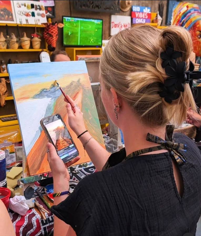 A visitor painting a mountain landscape on canvas at a Kitenge Arts class